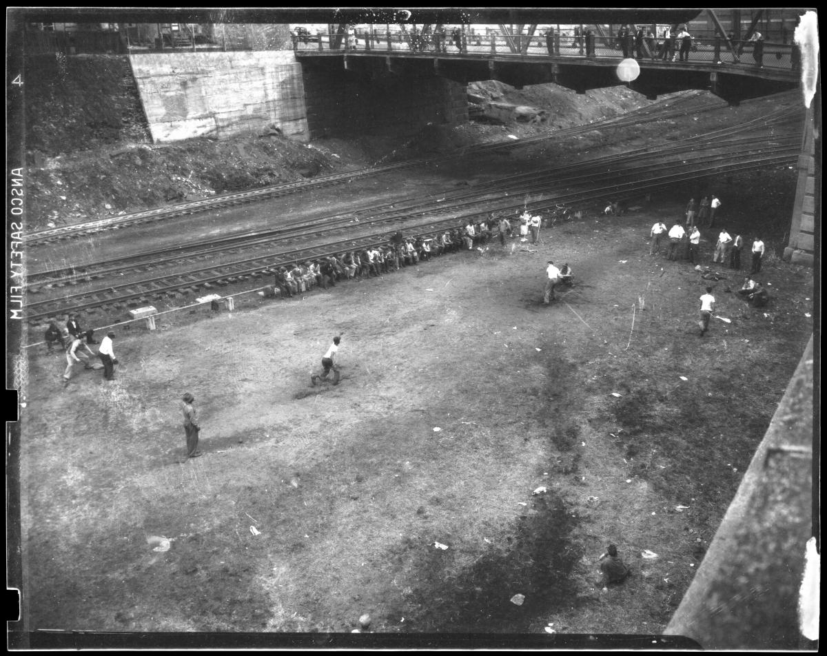 Overhead view of men playing a baseball game beside railroad tracks while spectators watch from the sidelines and a bridge above.
