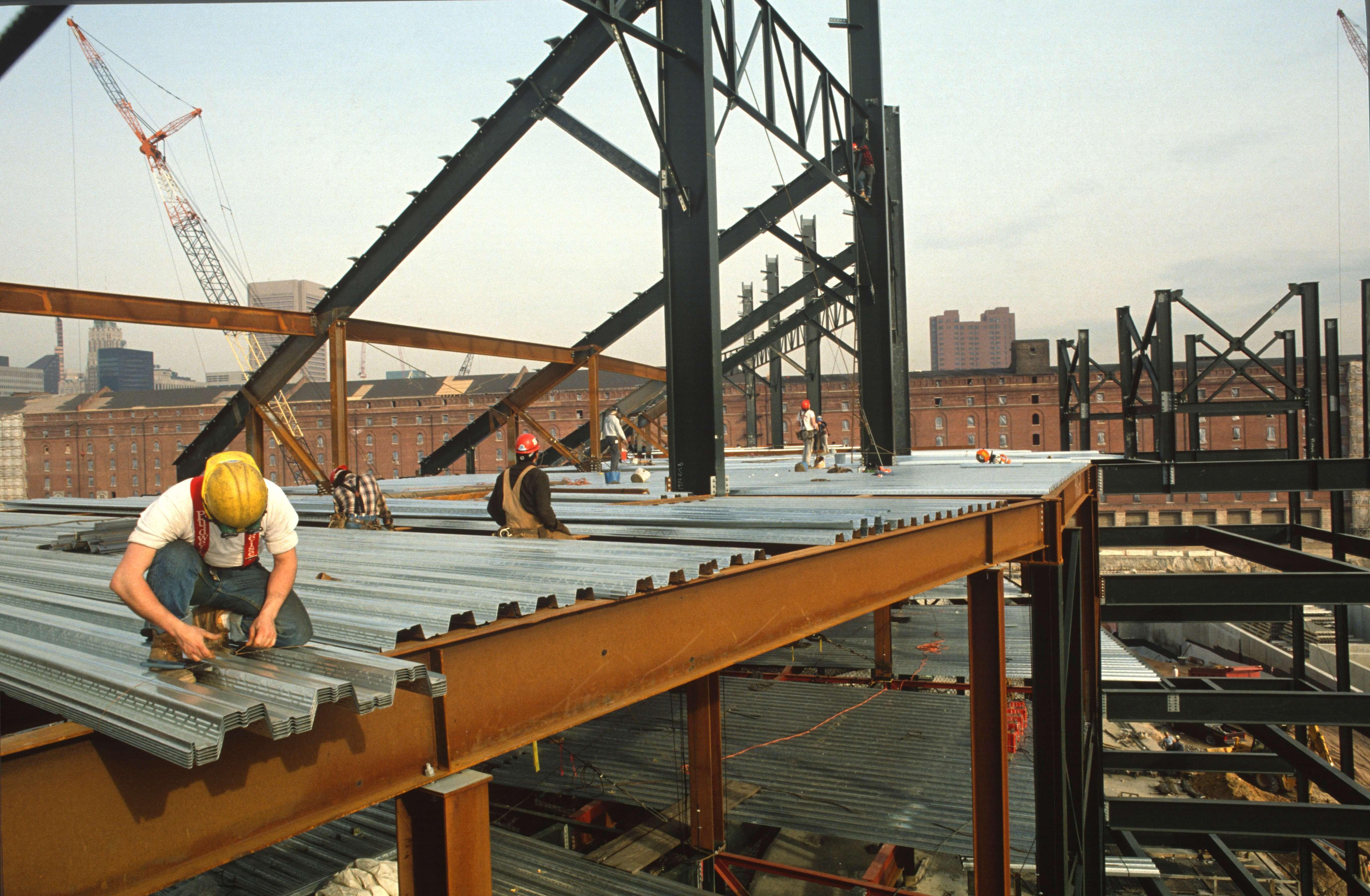 Workers installing metal decking on the steel skeleton of the stadium with the historic B&O Warehouse nearby.