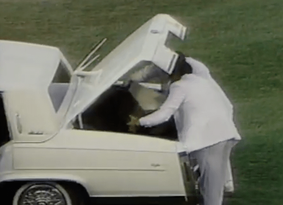 Two men in white suits place home plate into the trunk of a white car