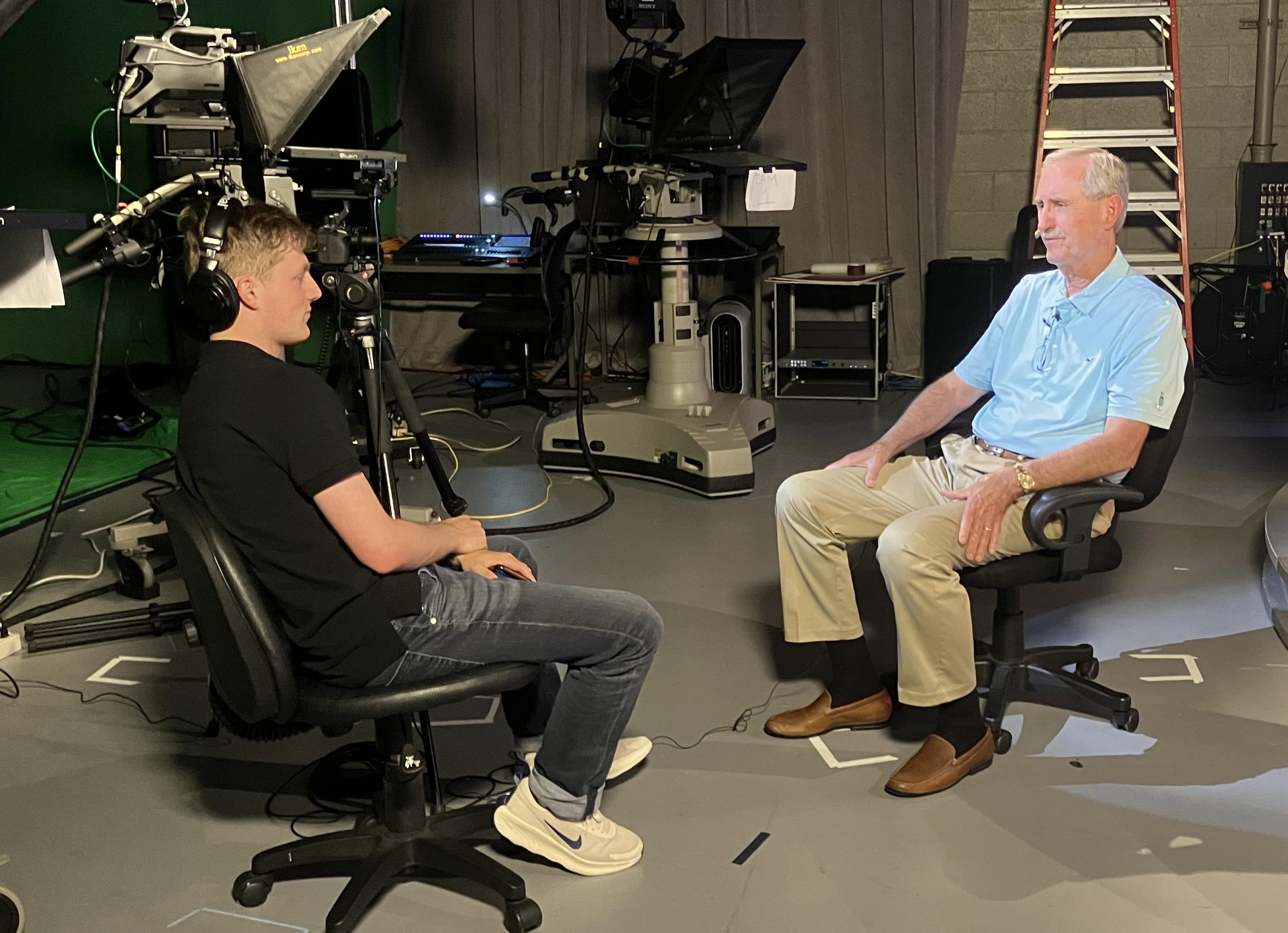 Two men seated facing each other during a filmed interview in a studio with cameras and lights.