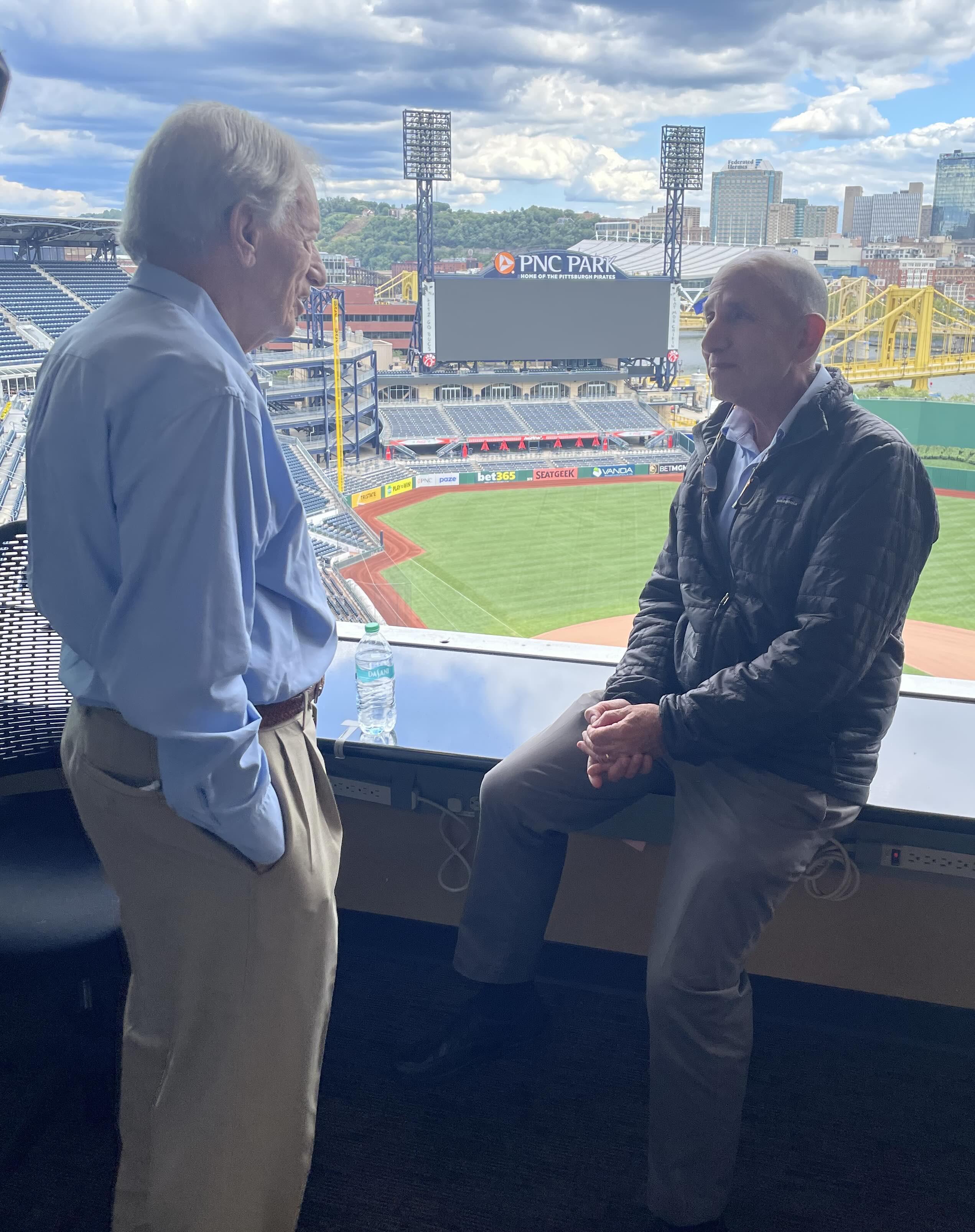 Two men talking in a press box overlooking a baseball field and stadium scoreboard.