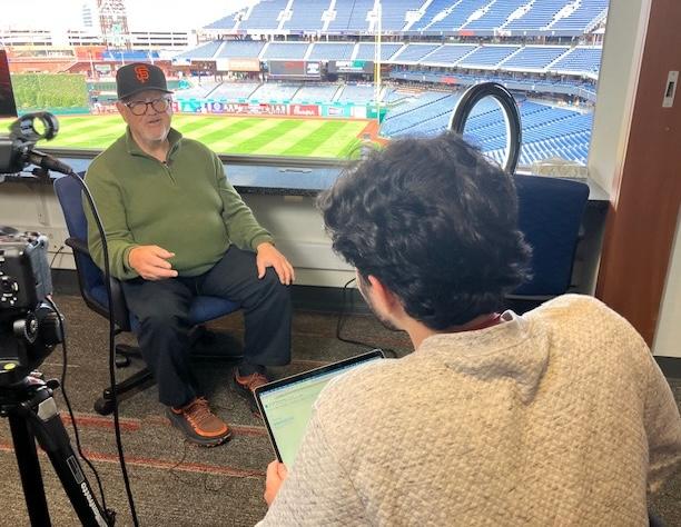 Man speaking during an interview while a camera, laptop, and ring light are set up nearby.