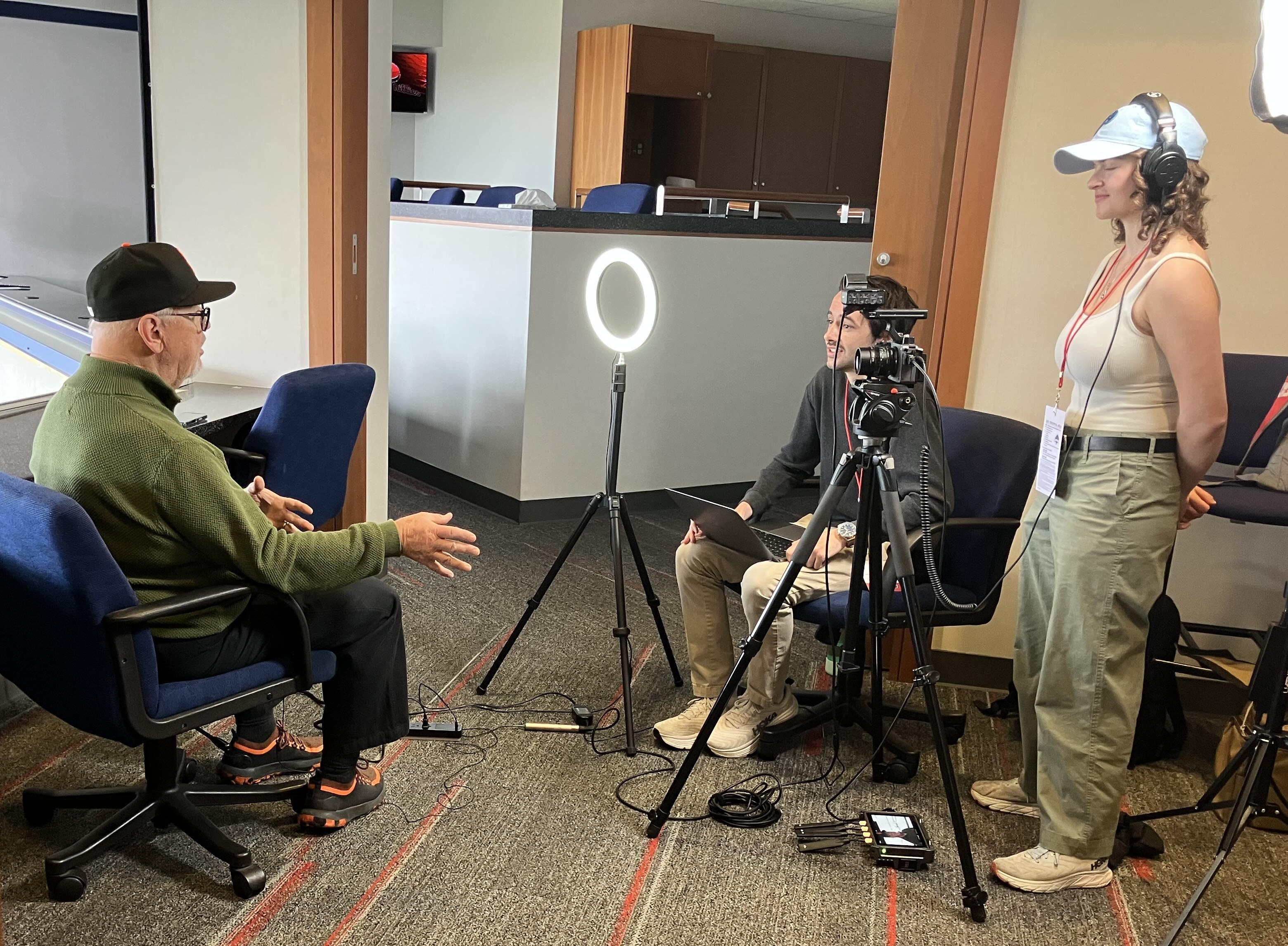 Man seated being interviewed by two people with a camera.