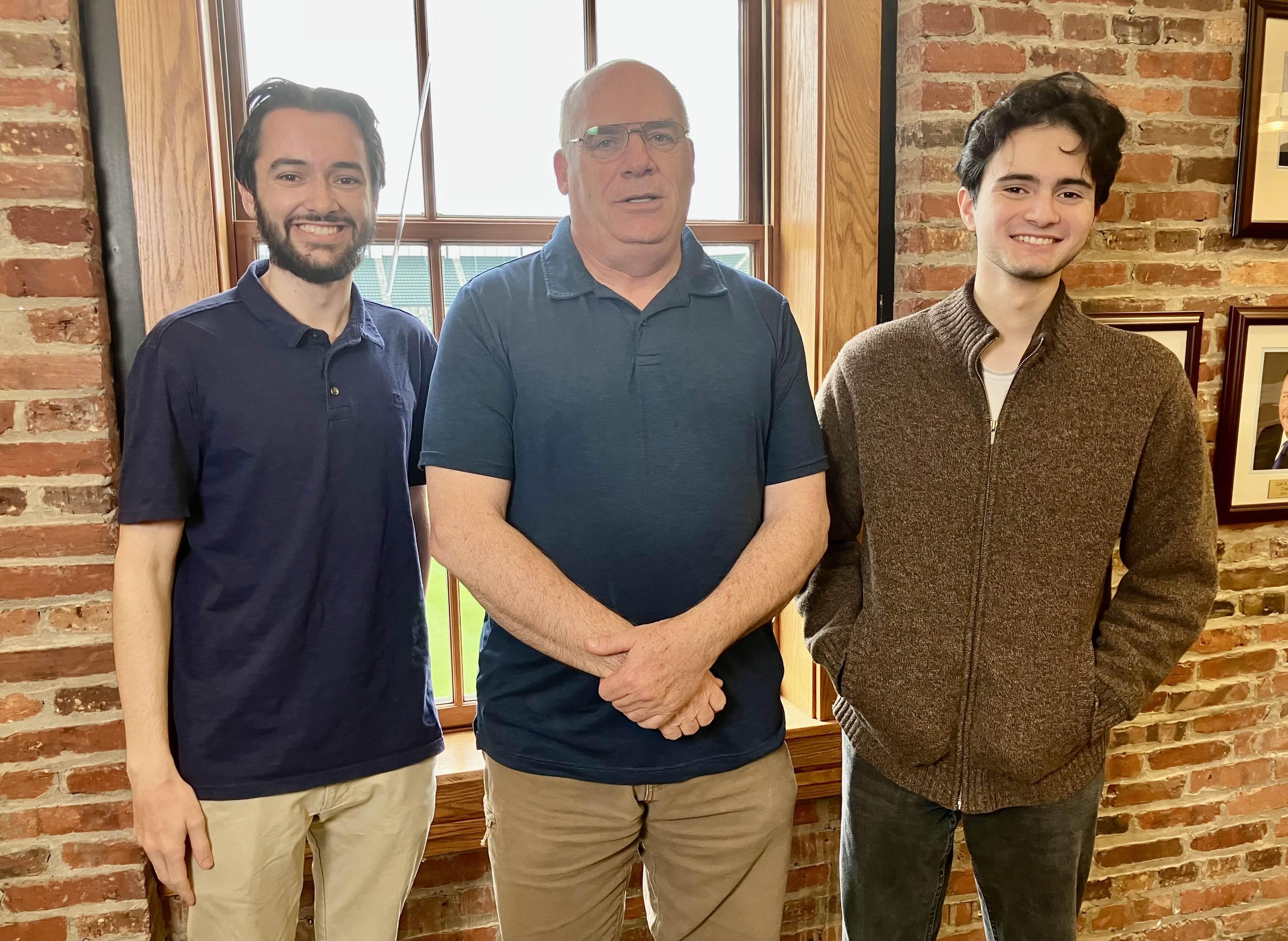 Three men standing together in front of a window and brick wall posing for a photo.