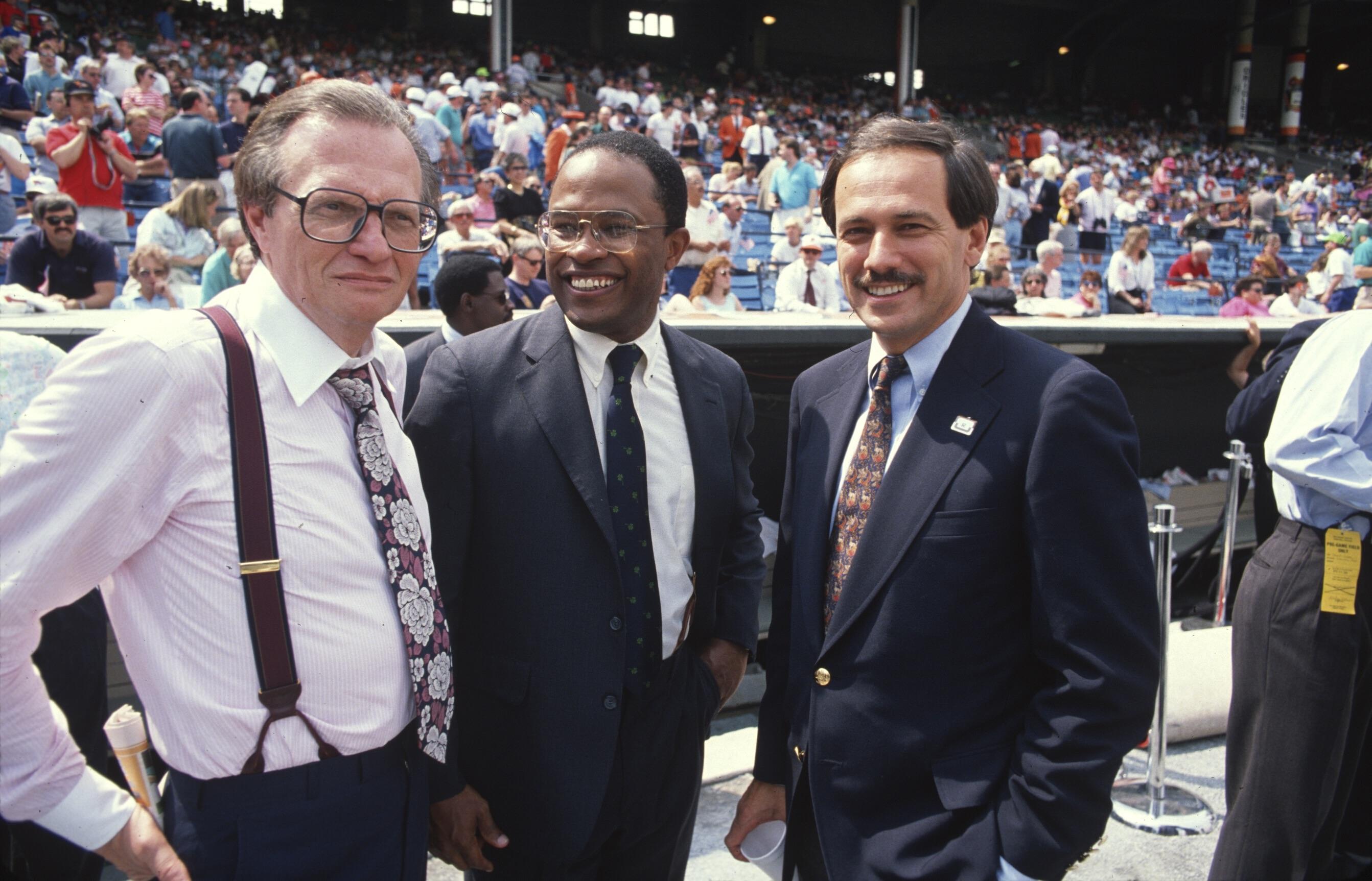 Three men in suits stand together on the field with a crowd of fans filling the stadium behind them.