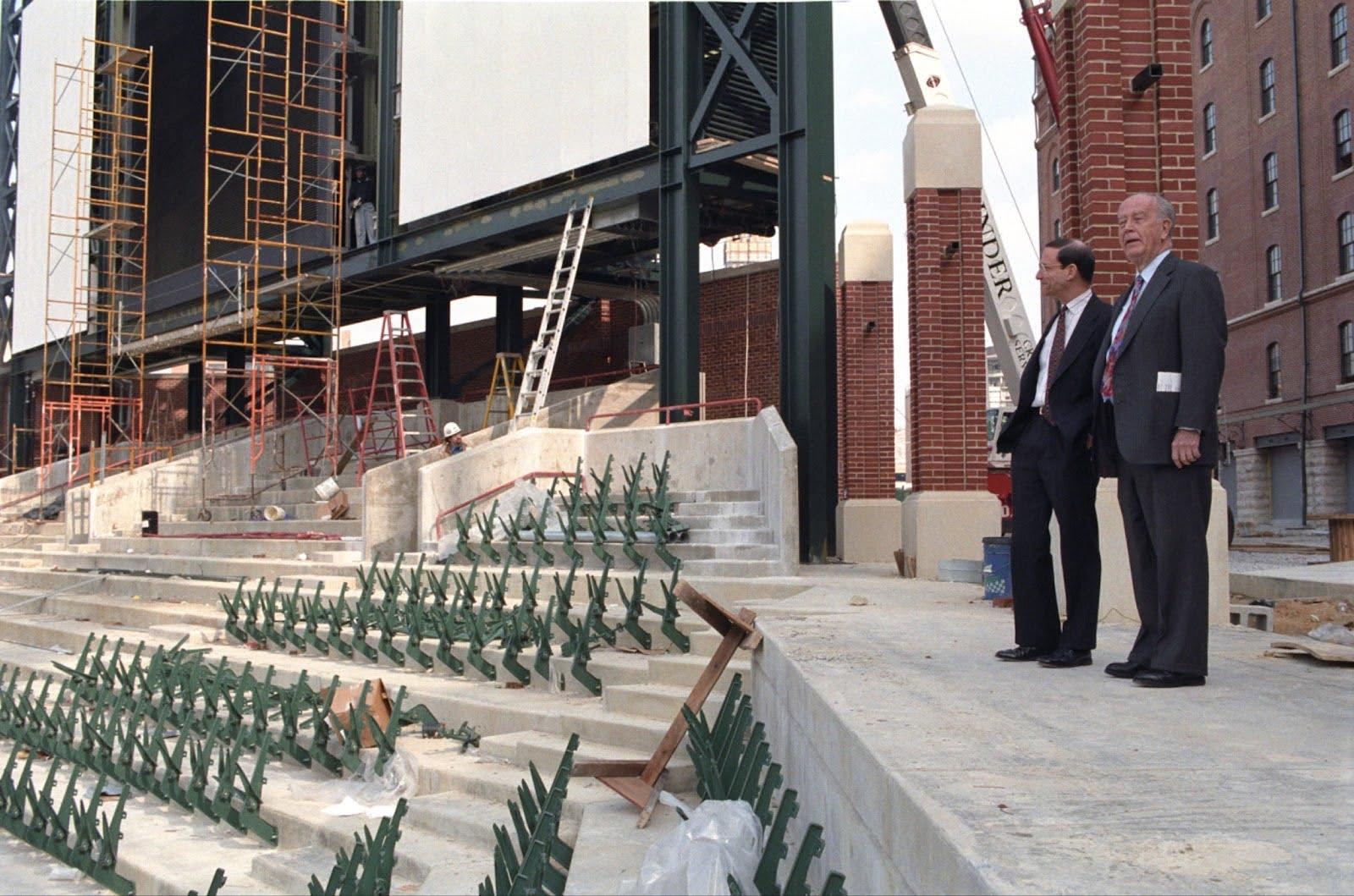 Officials observe construction progress at Oriole Park at Camden Yards with unfinished seating and structural steel visible.